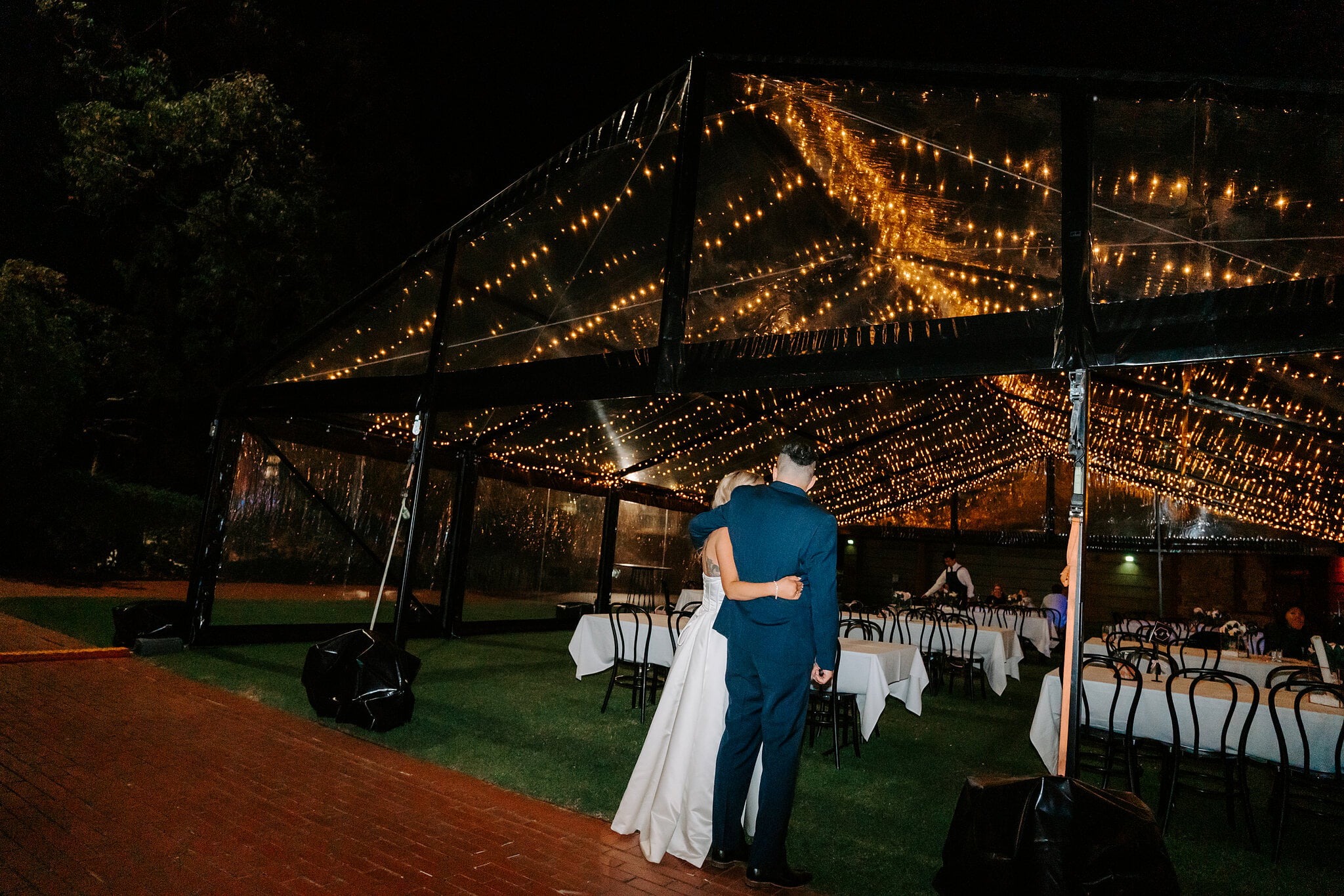 Sarah and Joe hold each other under the fairy-lit clear marquee at night.