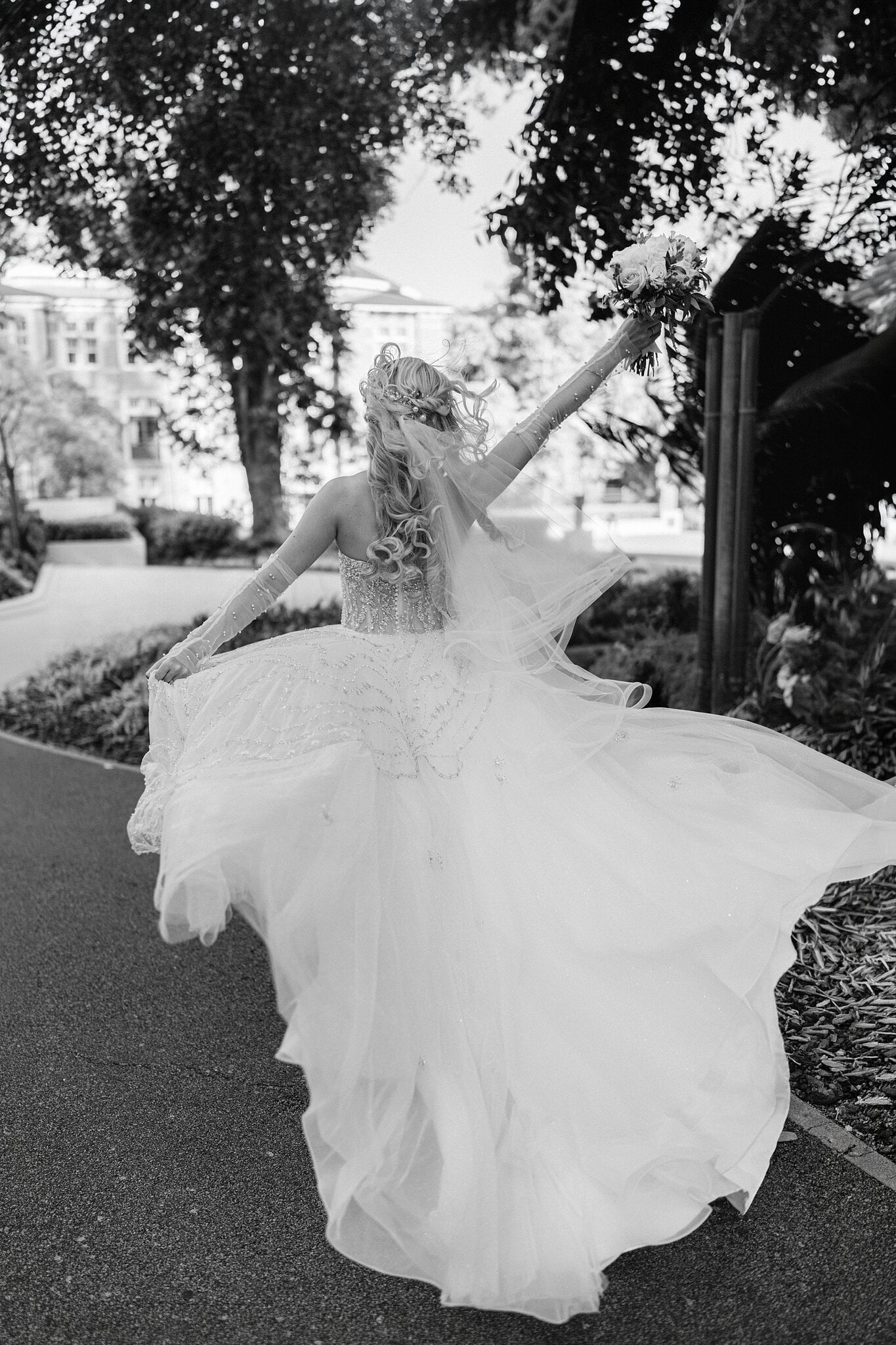 Black and white of Claire twirling her skirt with her bouquet raised under the old trees.
