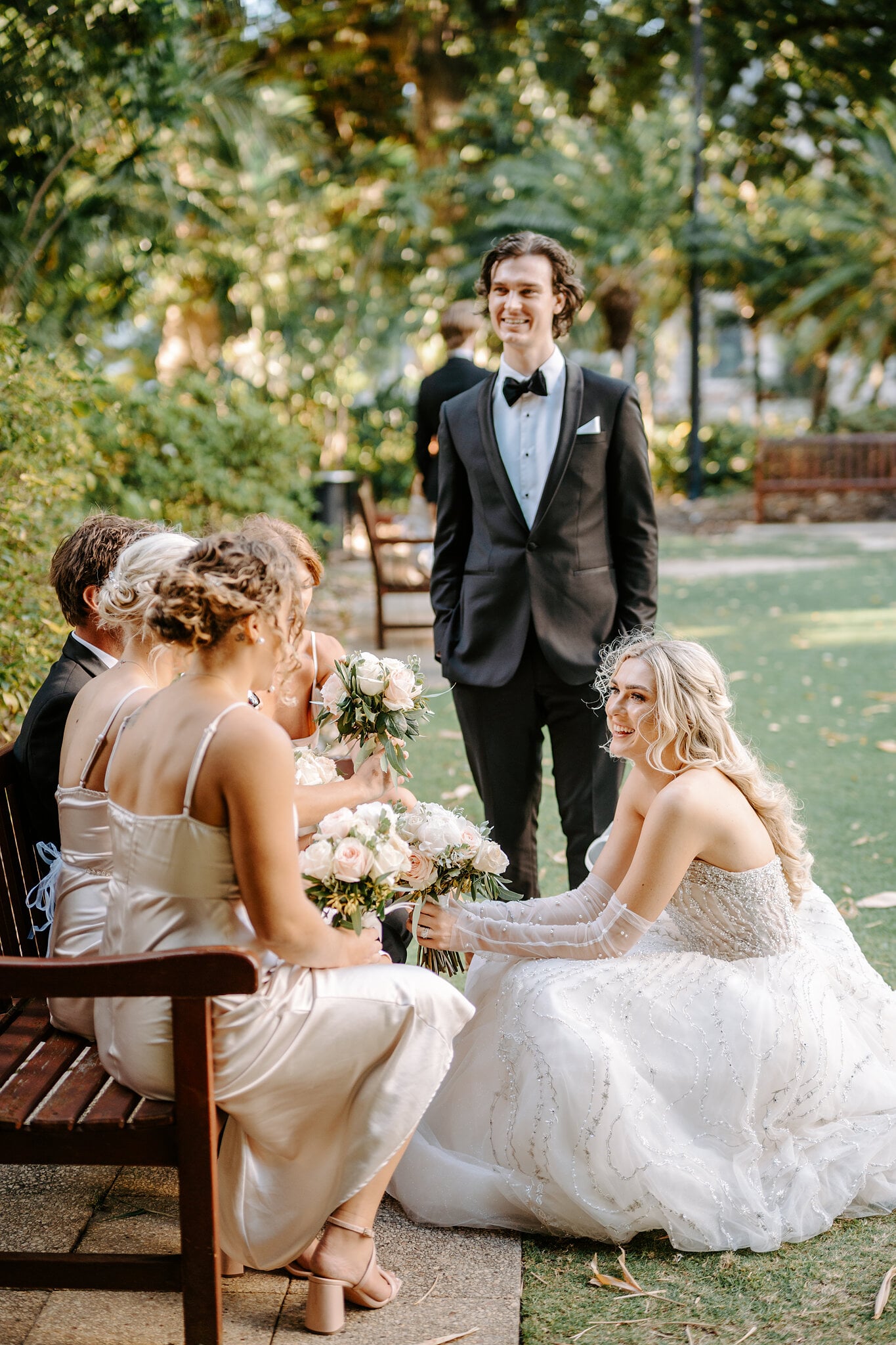 Claire crouches to chat with her bridesmaids on a garden bench as George smiles nearby.