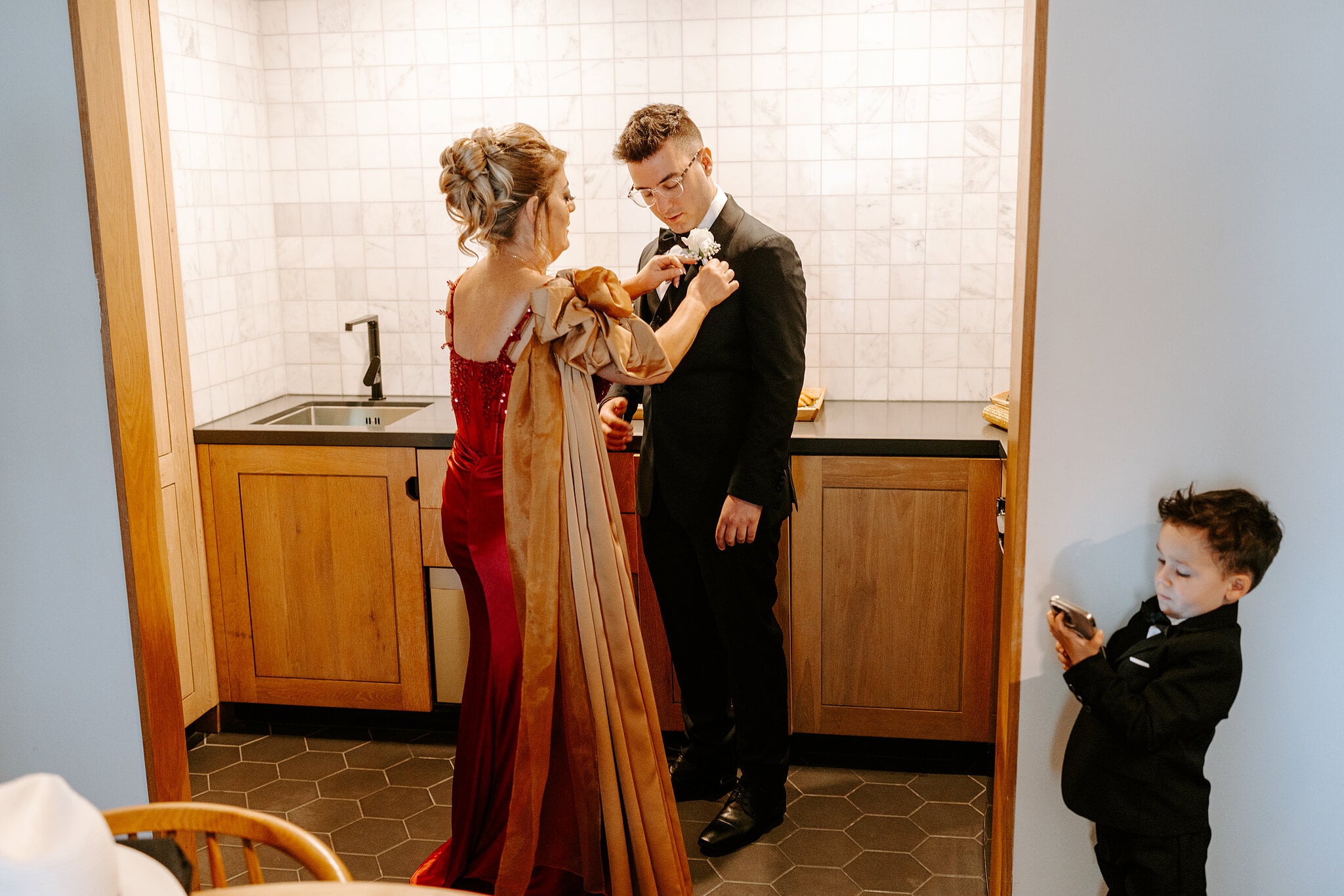 Jordan’s mum pins his buttonhole in his room at The Island while the littlest groomsman checks a phone in the corner.