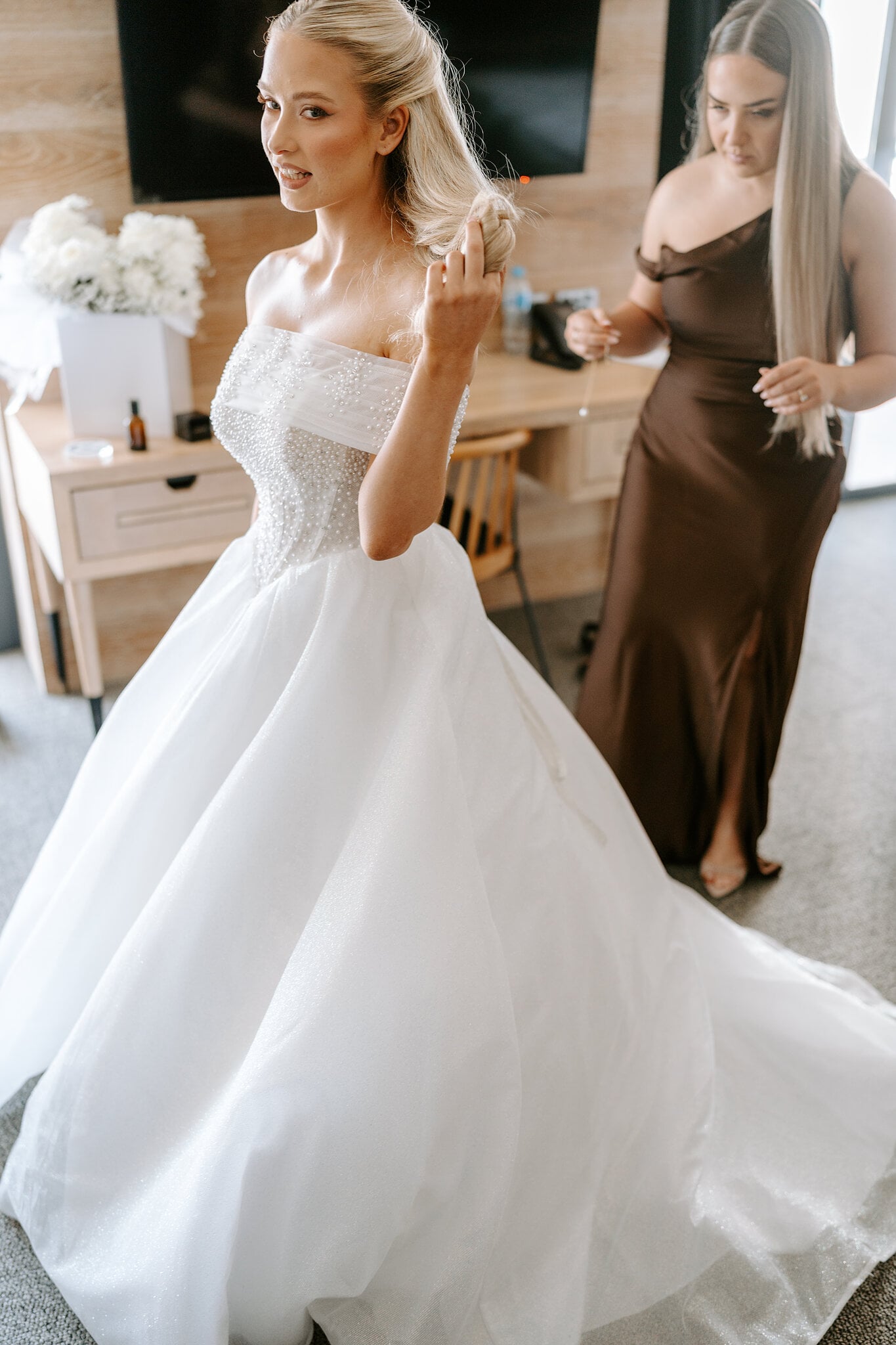 Sorayaa adjusts an earring, her full skirt spread across the floor as a bridesmaid in chocolate satin looks on.