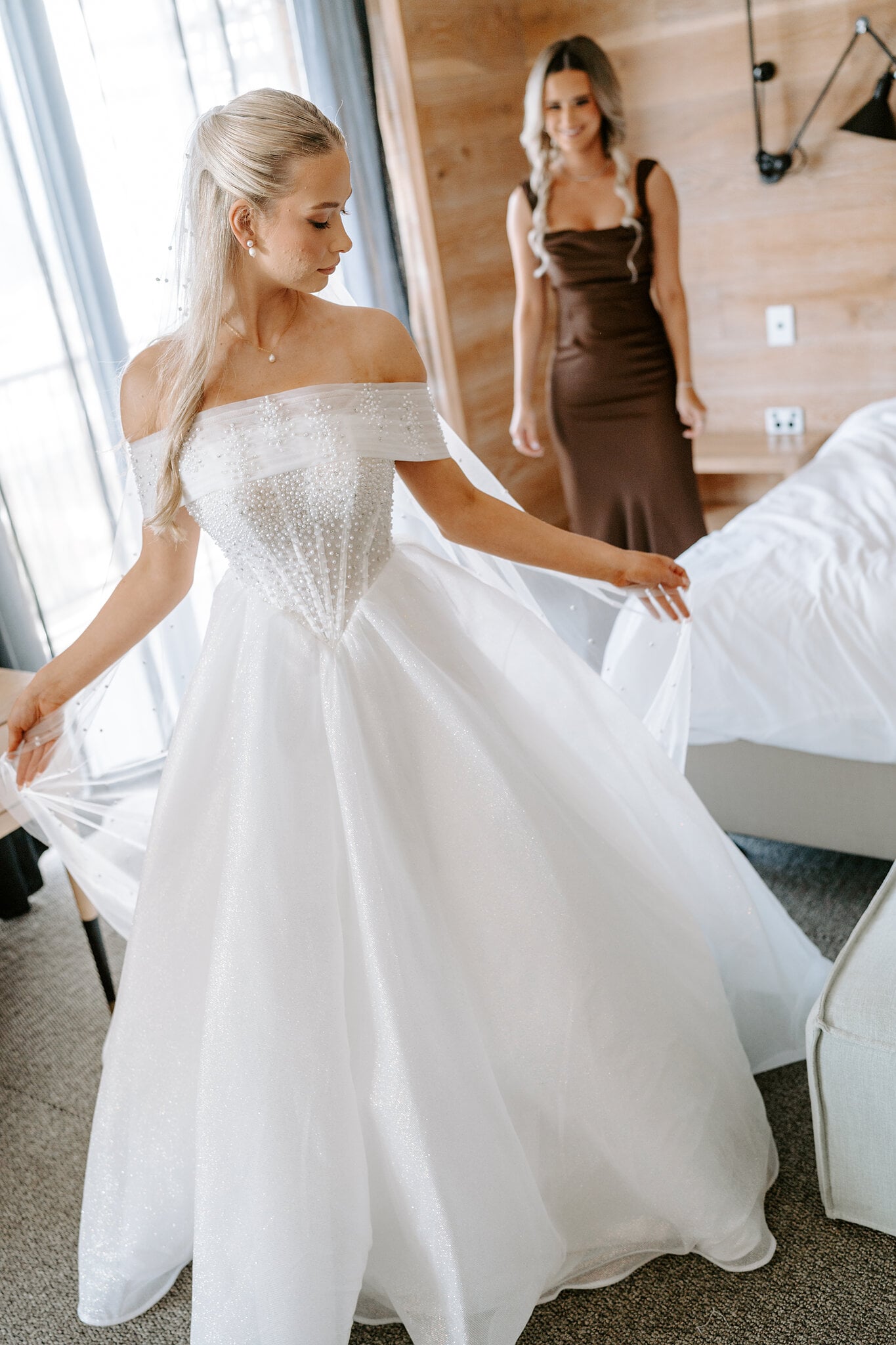 Sorayaa sweeps out her skirt in the window light while a bridesmaid watches.