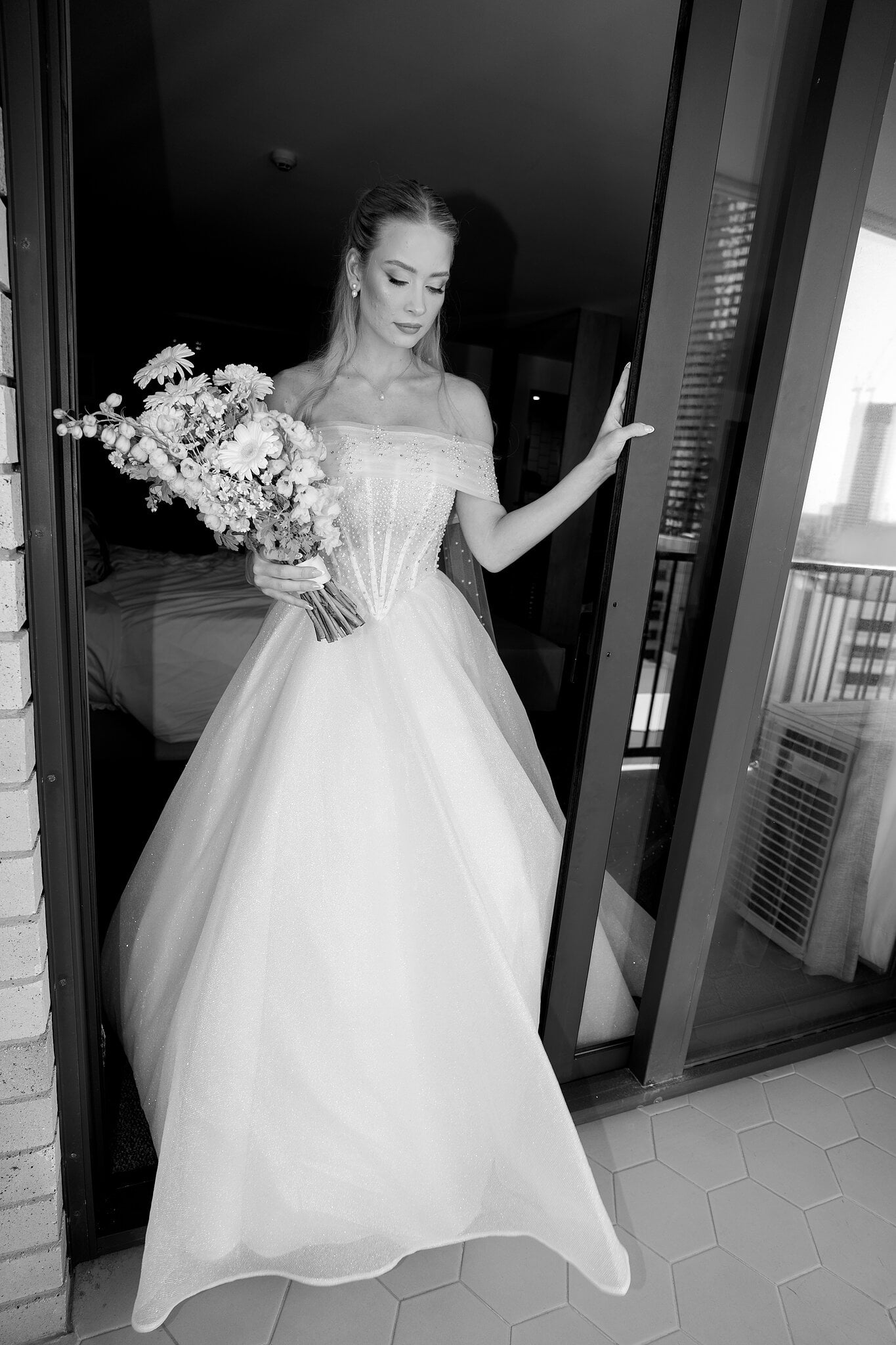 Sorayaa pauses in the balcony doorway with her bouquet, in black and white.