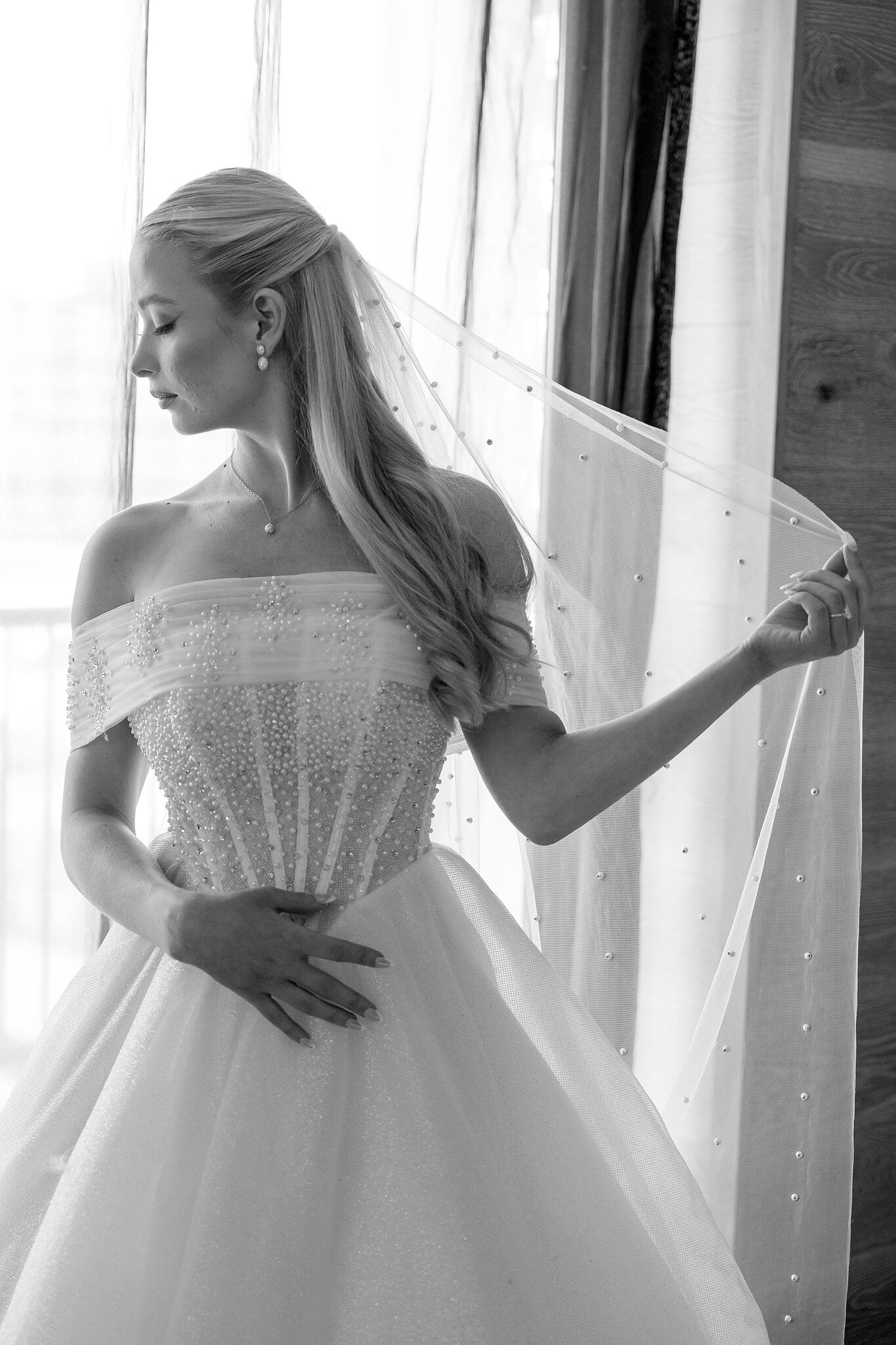 Sorayaa lifts her pearl veil by the window, in black and white.