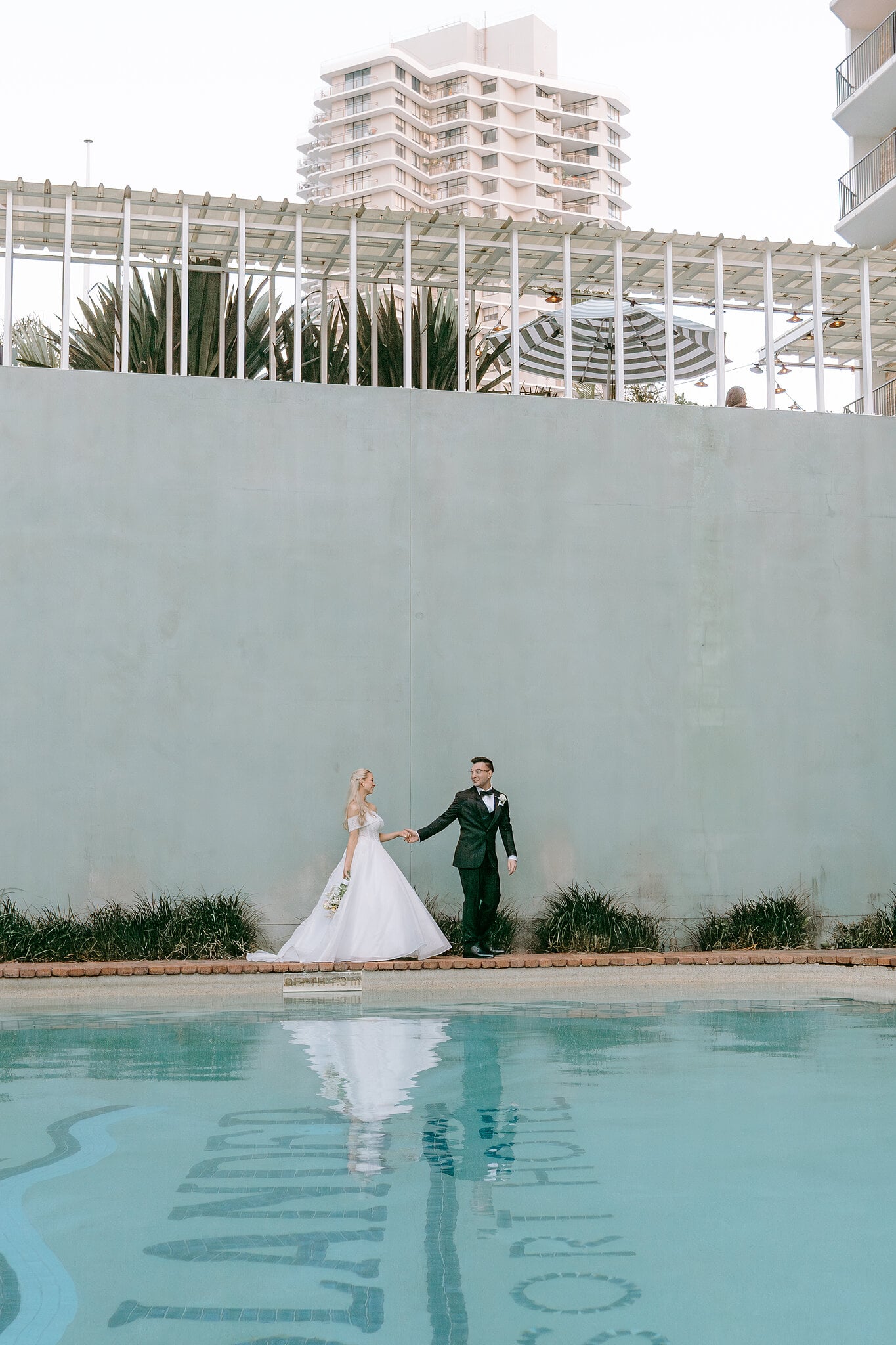 Sorayaa and Jordan stroll along the pool at The Island Gold Coast, reflected in the water above the old Islander logo.