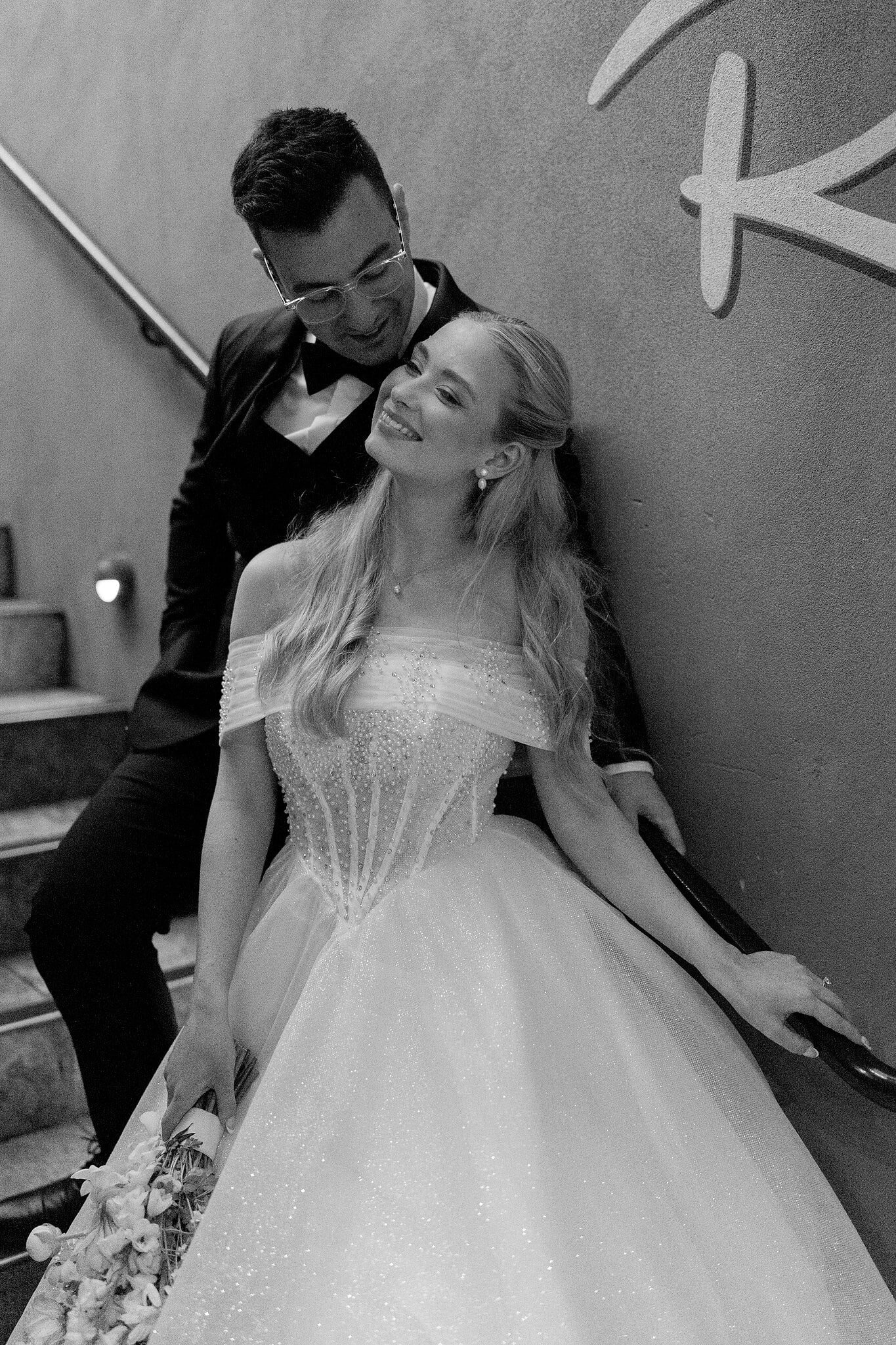 Sorayaa leans back into Jordan on the stairs, both laughing, in black and white.