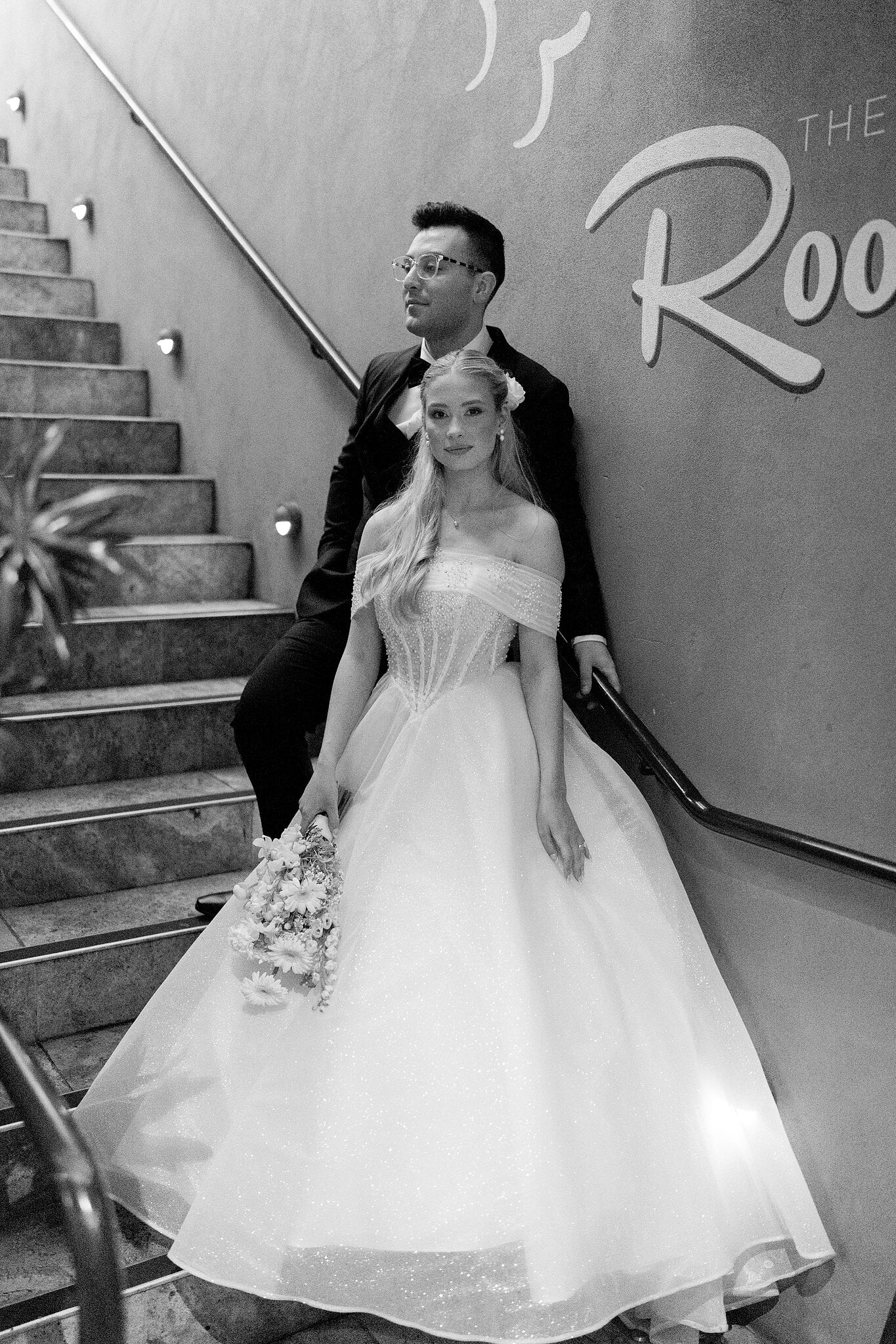 Sorayaa holds her bouquet on the stairs as Jordan looks out past her, in black and white.