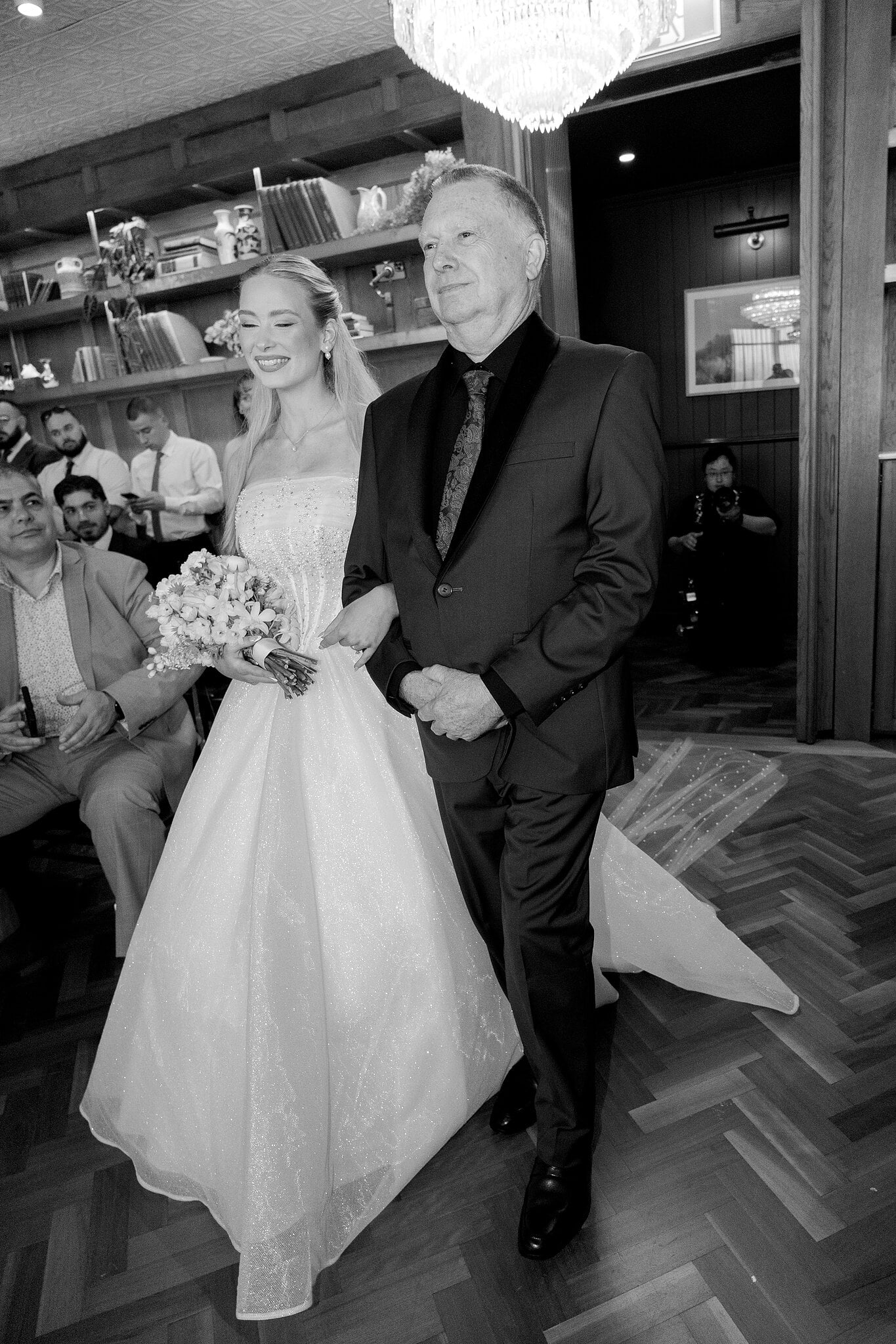Sorayaa walks down the aisle on her dad’s arm past the bookshelves of The Penthouse, in black and white.