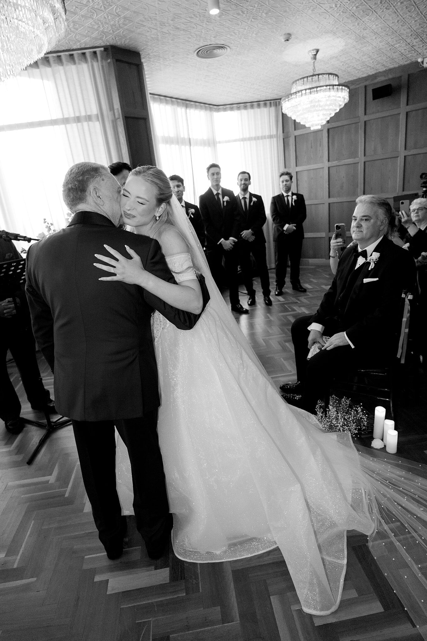 Sorayaa hugs her dad at the end of the aisle as the groomsmen look on, in black and white.