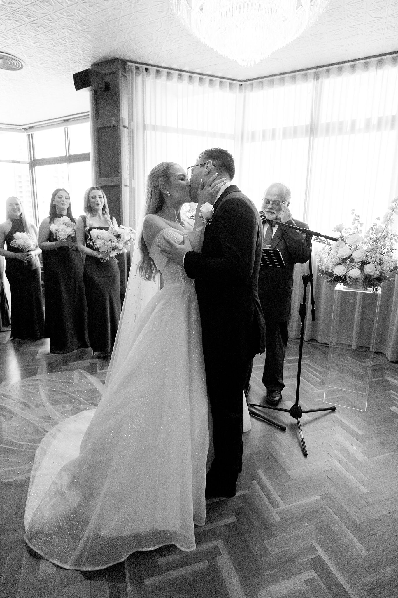 The first kiss in The Penthouse at The Island Gold Coast, bridesmaids lining the windows, in black and white.