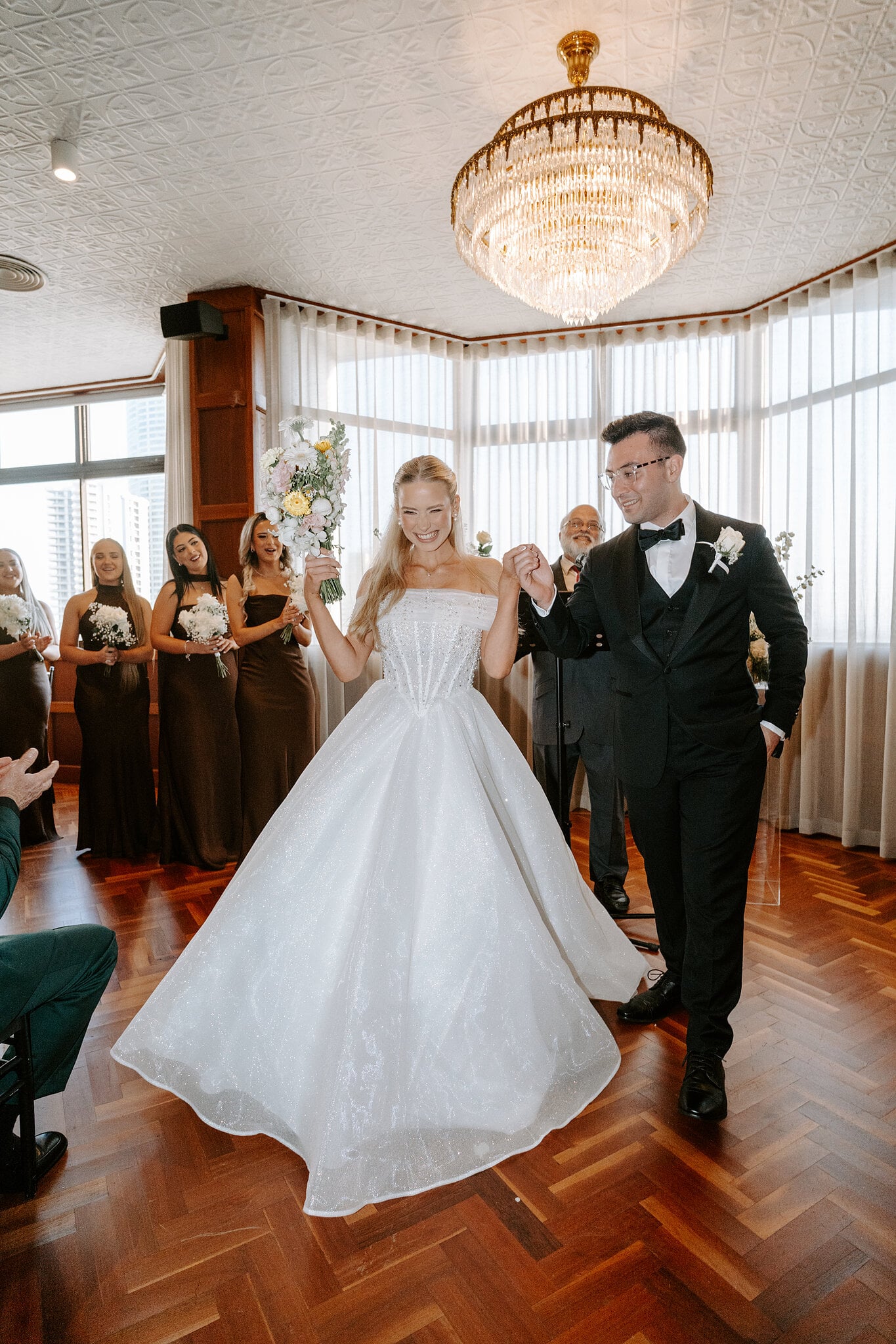 Just married, Sorayaa and Jordan head back up the aisle under the chandelier, her bouquet raised.