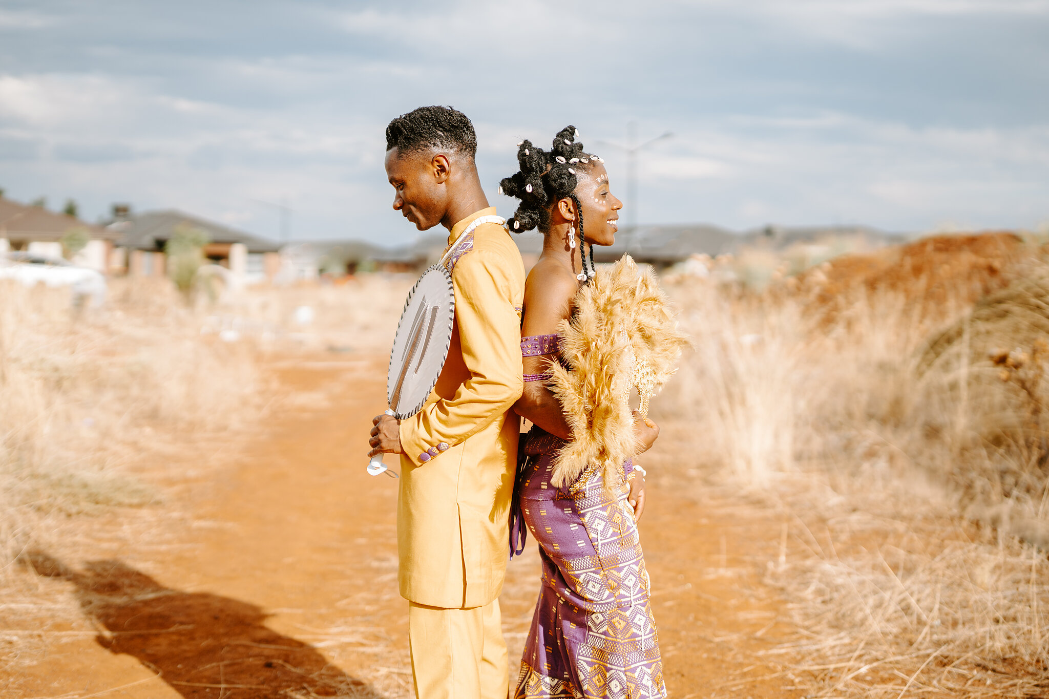 Allen and Pearl stand back to back in gold and purple traditional dress on a red dirt track.