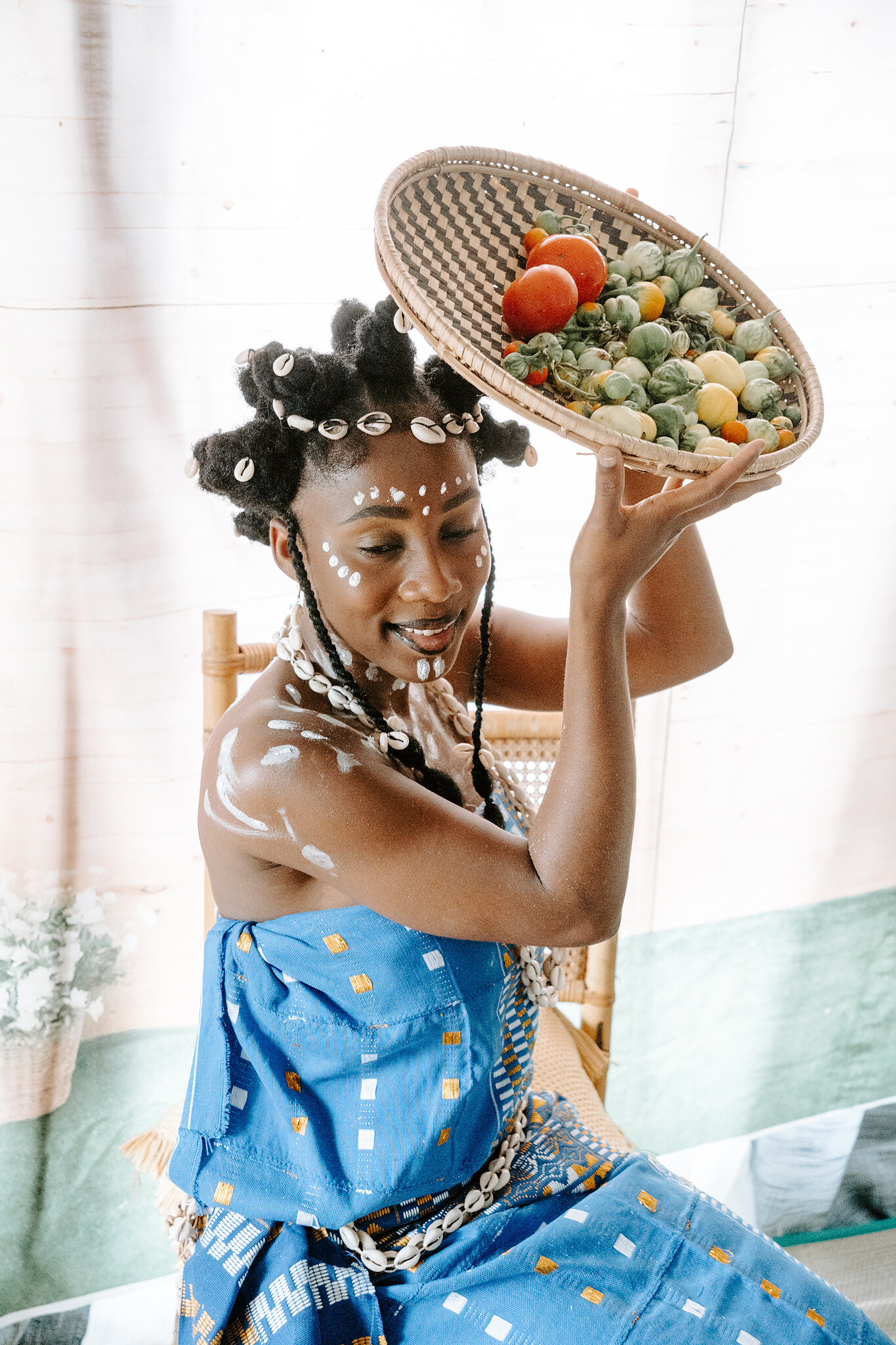 A maiden with white body markings and cowrie shells lifts a basket of fresh produce overhead.