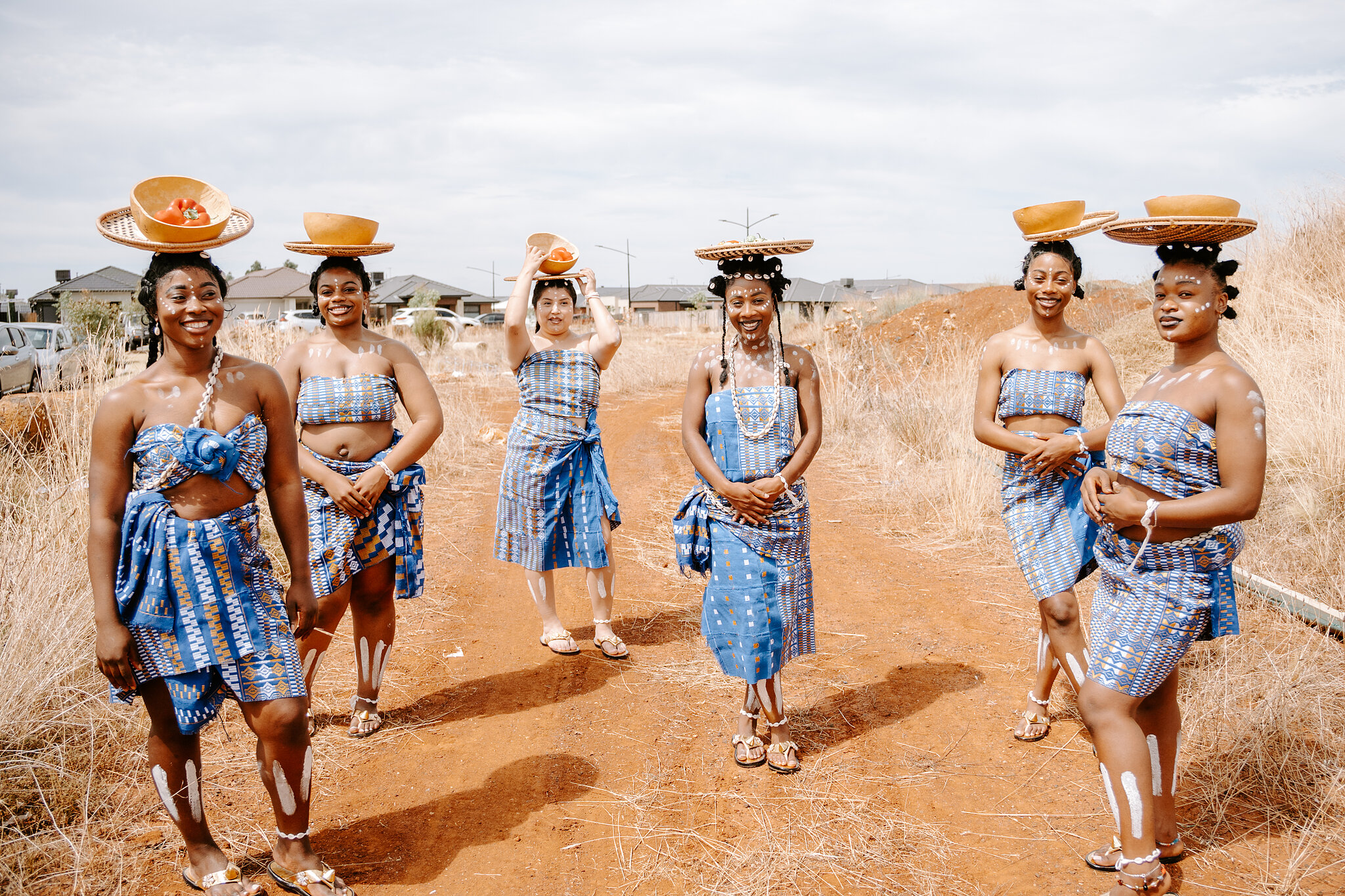 Six maidens in blue woven cloth carry baskets and calabash bowls on their heads along a dirt track.