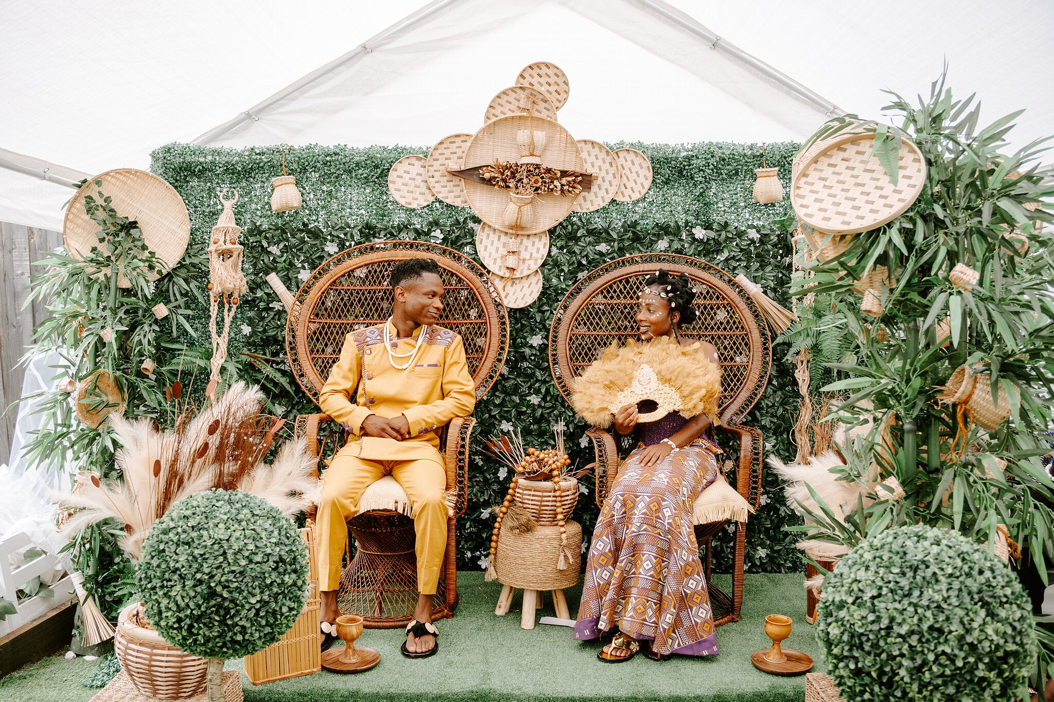 Pearl and Allen on rattan peacock chairs before a wall of greenery and woven baskets in the marquee.