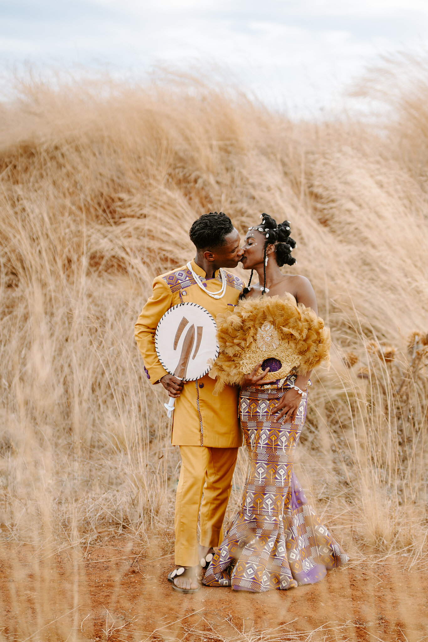 Pearl and Allen kiss in tall golden grass in their gold and purple traditional dress.